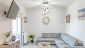 Un salon moderne avec un canapé d’angle gris, une table basse en bois, un téléviseur posé sur un meuble, une horloge murale et un ventilateur de plafond.