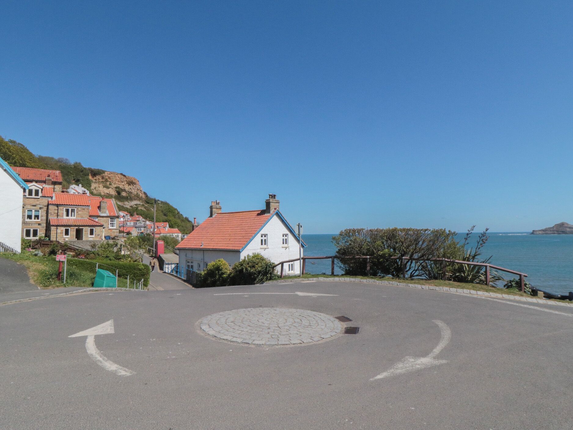 Eine Küstenstraße mit Kreisverkehr, Häuser mit roten Dächern und freiem Blick auf das Meer.