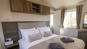 A neatly made bed with a grey headboard, a bedside table with a power strip, and a window with curtains. - Foxglove Lodge (Saltburn-by-the-Sea)