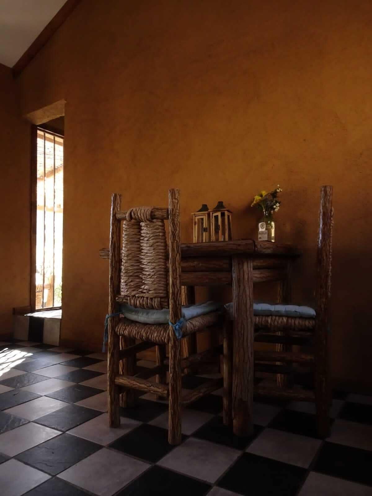 Una habitación con piso a cuadros, dos sillas de madera rústica con asientos de mimbre, una mesa de madera y una ventana con persianas verticales.
