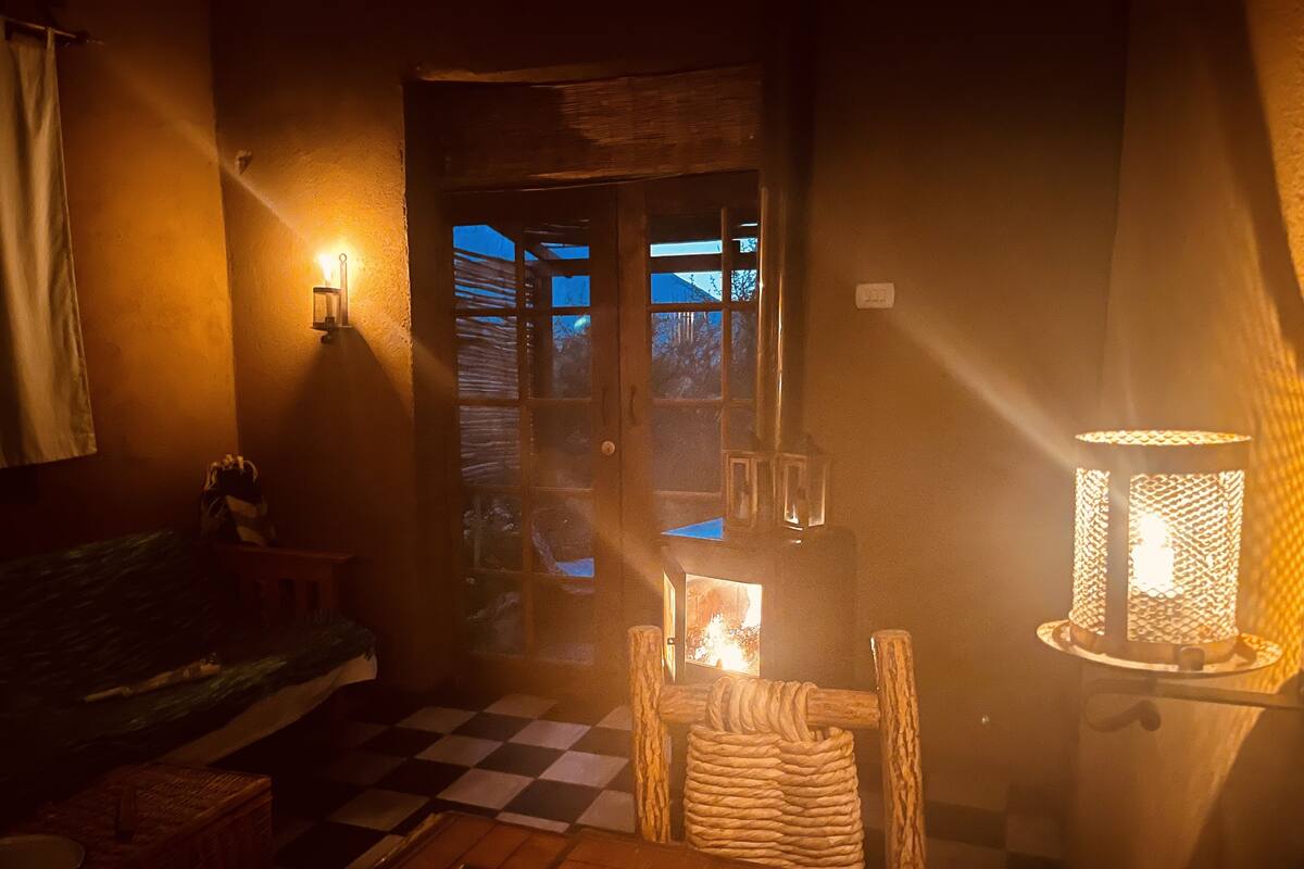 Habitació acollidora amb un paviment a quadres, una taula de fusta, una cistella teixida, una làmpada encèsa i una vista de l'exterior per una porta.