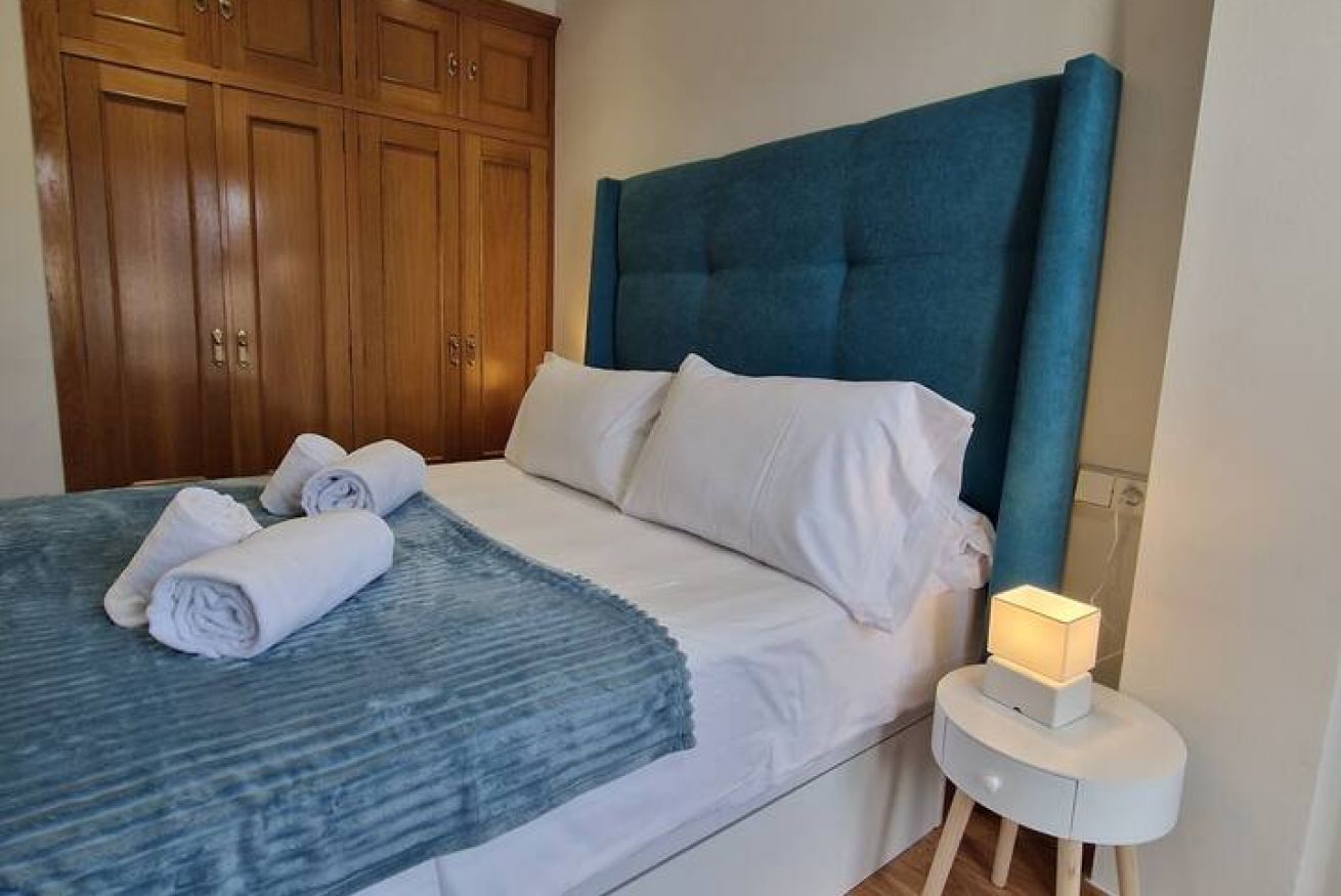 Una cama bien tendida con sábanas blancas y un cabecero azul, un armario de madera y una mesita de noche con una lámpara.
