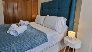 Uma cama bem arrumada com roupa de cama branca e cabeceira azul, um guarda-roupa de madeira e uma mesa de cabeceira com um abajur.