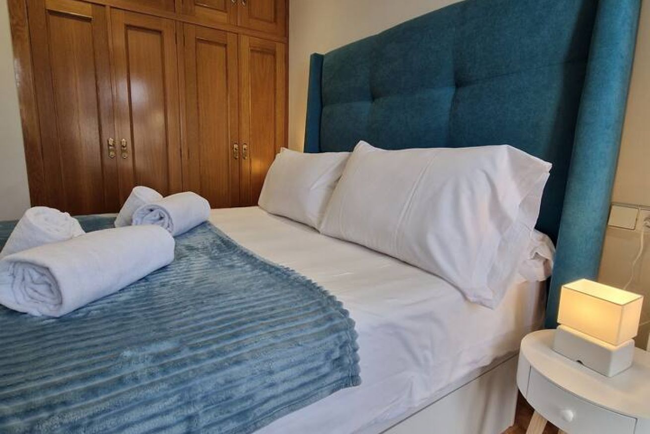 Una cama bien hecha con sábanas blancas y un cabecero azul, un armario de madera y una lámpara de noche.