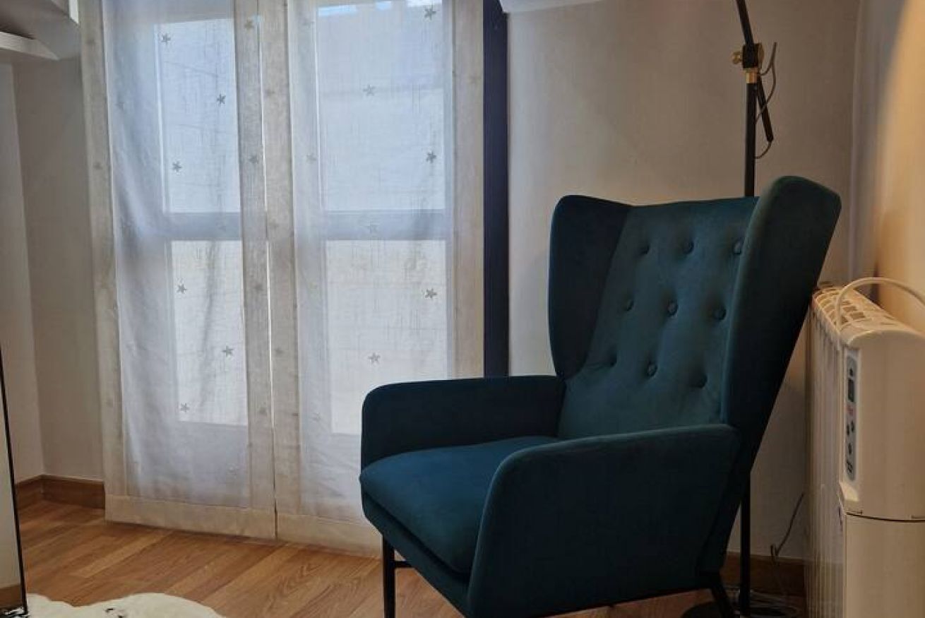 Een kamer met een blauwe beklede stoel, een raam met dunne gordijnen en een witte radiator.
