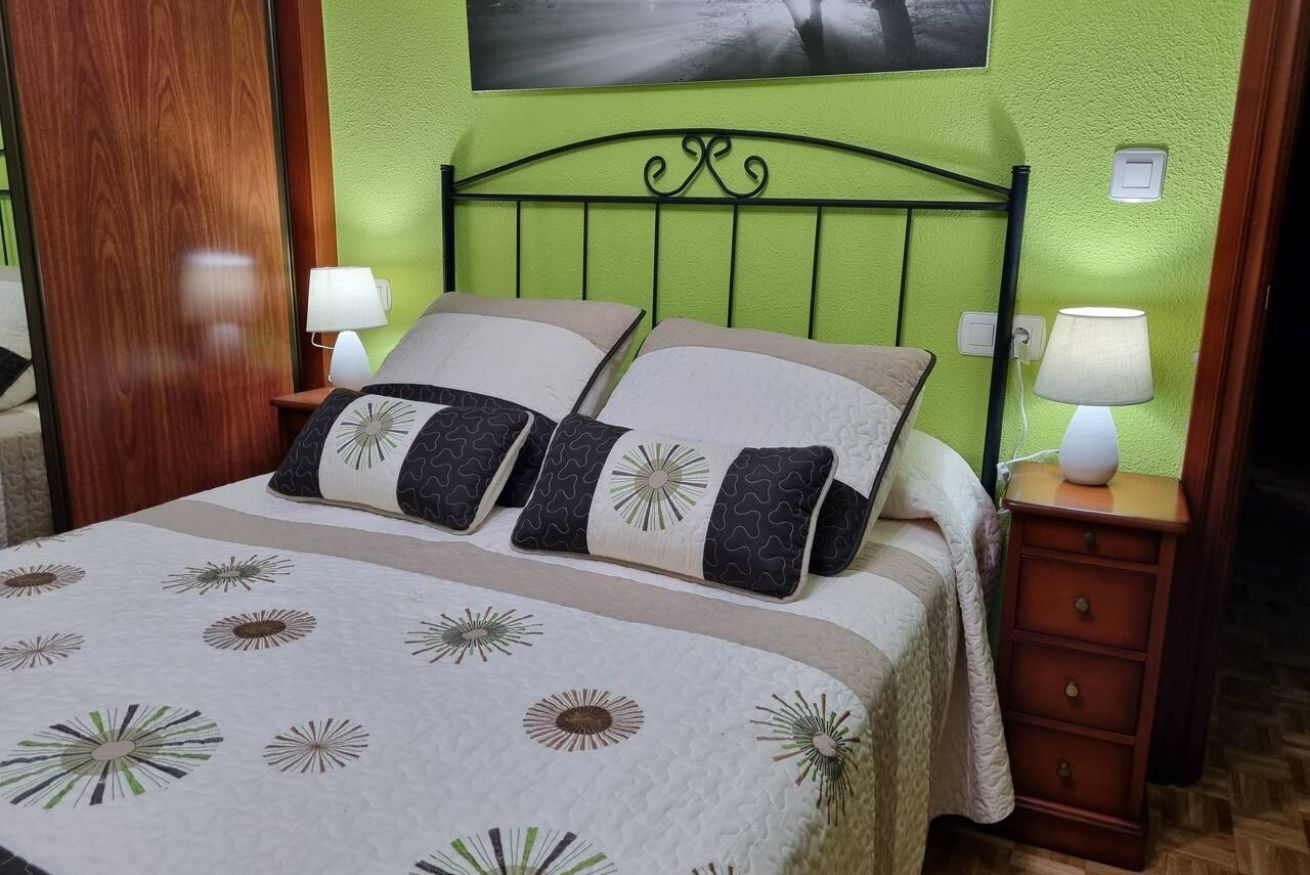 A bed with a floral-patterned bedspread, two pillows with a geometric design, a green wall, and a wooden nightstand with two lamps.