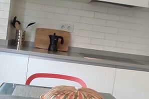 Una cocina con armarios blancos, una silla roja, una tabla de cortar de madera y una cafetera.