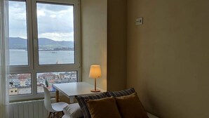 Una habitación con cama, una mesa, una silla y una lámpara. Hay ventanas con vistas a un cuerpo de agua y edificios.