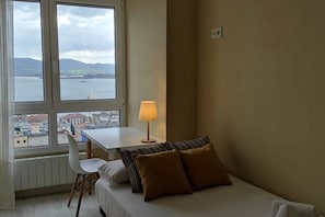 Una habitación con cama, una mesa, una silla y una lámpara. Hay ventanas con vistas a un cuerpo de agua y edificios.