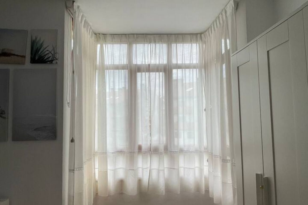 Una habitación con un ventanal cubierto por cortinas translúcidas, una puerta a la derecha y obras de arte colgadas en la pared.