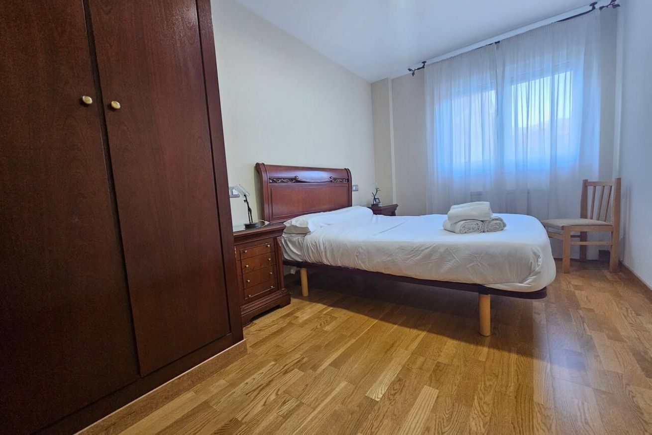 Un dormitorio con un armario de madera, una cama con colchón blanco y una toalla enrollada, una mesita de noche de madera, y una silla.