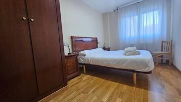 Un dormitorio con un armario de madera, una cama con colchón blanco y una toalla enrollada, una mesita de noche de madera, y una silla.