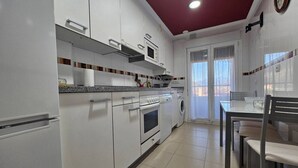 A kitchen with white cabinets, a microwave, oven, and a washing machine. There is a dining table with chairs and a window with curtains.