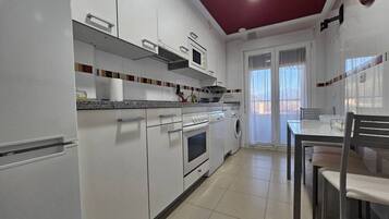 Een keuken met witte kasten, een magnetron, oven en een wasmachine. Er staat een eettafel met stoelen en een raam met gordijnen.