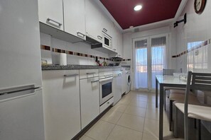 A kitchen with white cabinets, a microwave, oven, and a washing machine. There is a dining table with chairs and a window with curtains. - 22A02 Modern Apartment in Arriondas by R2R Consulting (Arriondas)