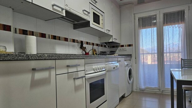 A modern kitchen with white cabinets, a built-in oven, a washing machine, and a dining area with a table and chairs.
