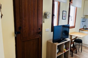 A room with a wooden door, a television on a wooden stand, a kitchen area with a window, and a small stool.