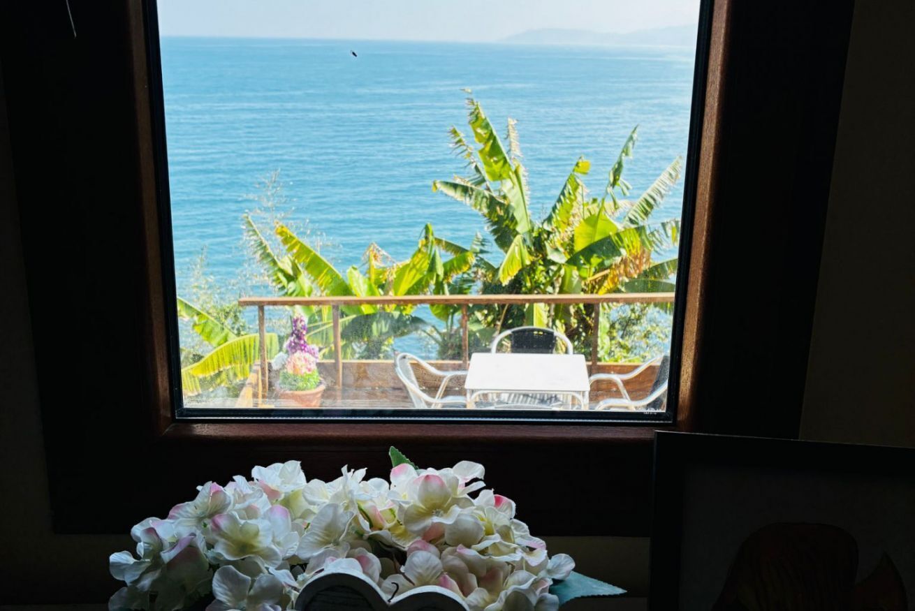 Una habitación con vista al océano a través de una ventana, una planta en maceta y una mesa con sillas.