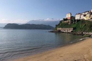 A coastal beach with buildings on a cliff, calm water, and distant mountains. - The LLastrina 14A03 by R2R Consulting (Lastres)
