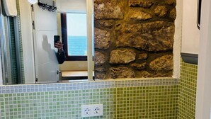 Casa de banho com azulejos de mosaico verde, parede de pedra, espelho e vista para o mar.