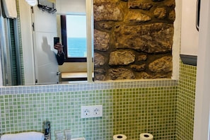 A bathroom with green mosaic tiles, a stone wall, a mirror, and a view of the sea.