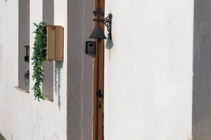 A white building with a wooden door, a black wall-mounted light, and a wooden planter with green plants. - The LLastrina 14A03 by R2R Consulting (Lastres)