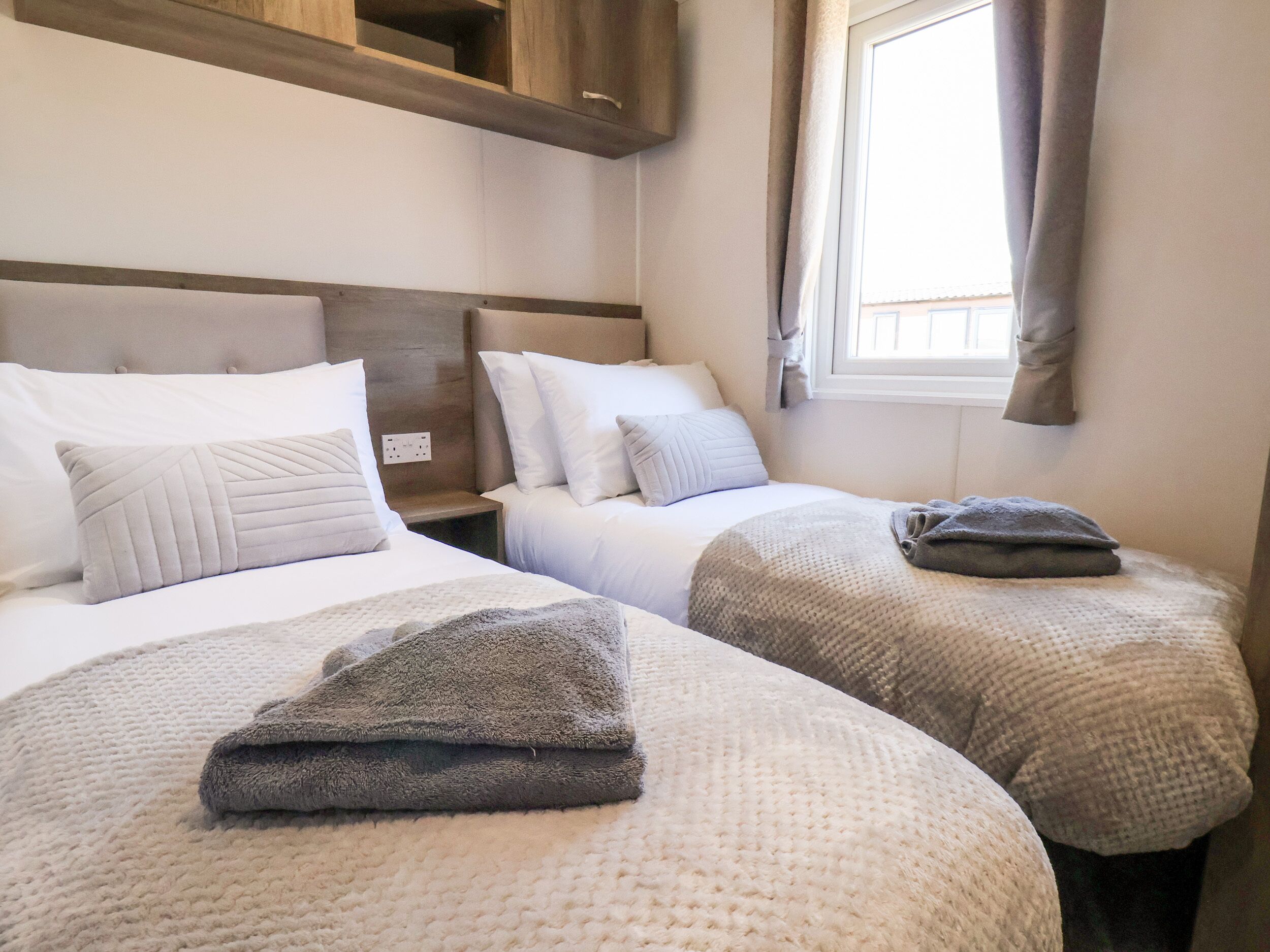 A hotel room with two beds, each with white bedding and a grey blanket folded on top. There is a window with curtains, a wooden headboard, and a wall socket visible.