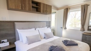 Een net opgemaakt bed met een grijze deken en kussens, een houten kledingkast erboven en een raam met gordijnen.