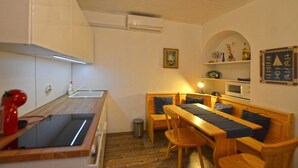 Cocina compacta con cubierta de madera, un lavamanos y una tetera roja. Hay una zona de comedor con una mesa de madera y bancos, y un microondas en la pared.