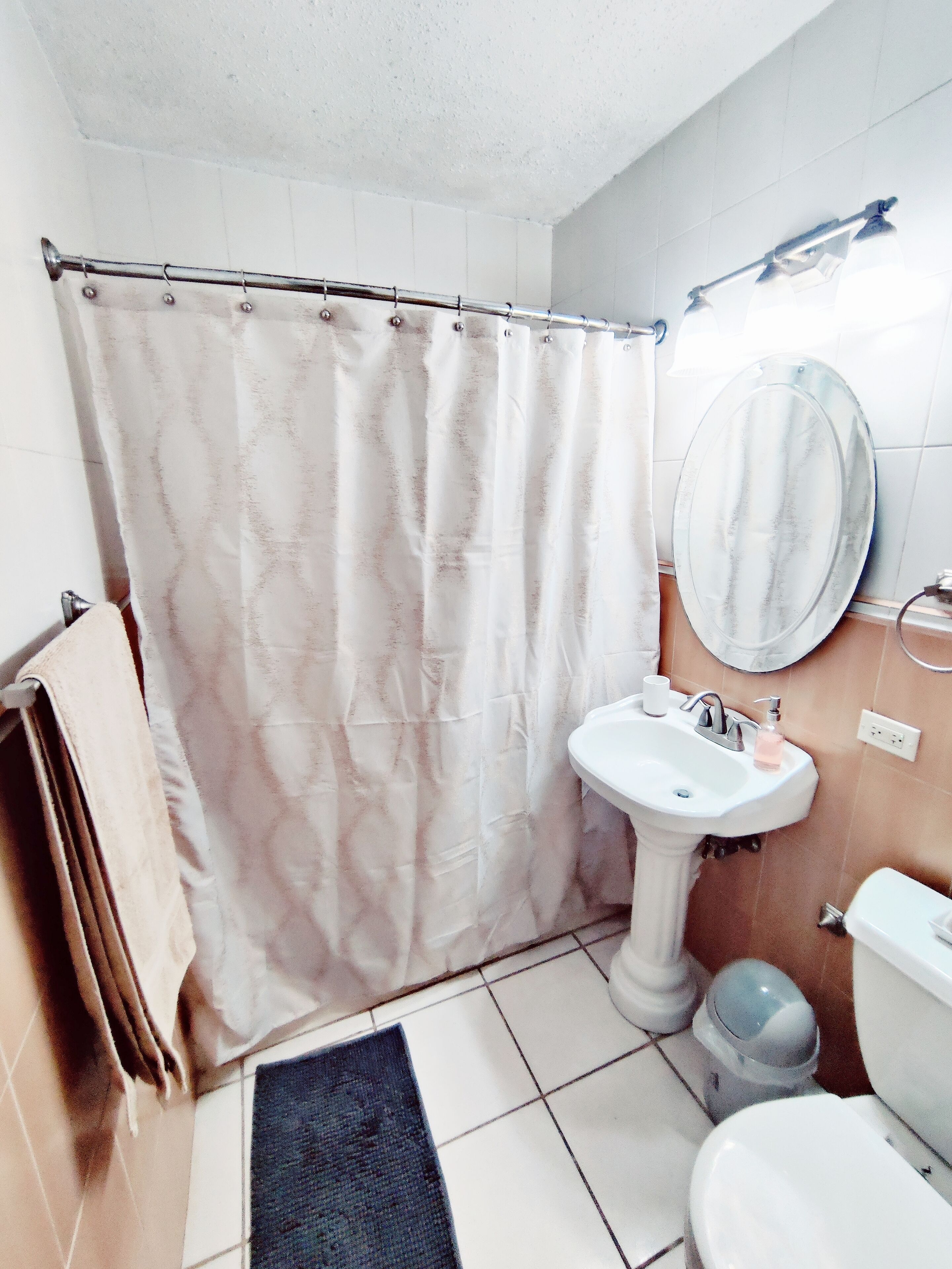 A bathroom with a shower curtain, sink, mirror, toilet, and a trash bin.