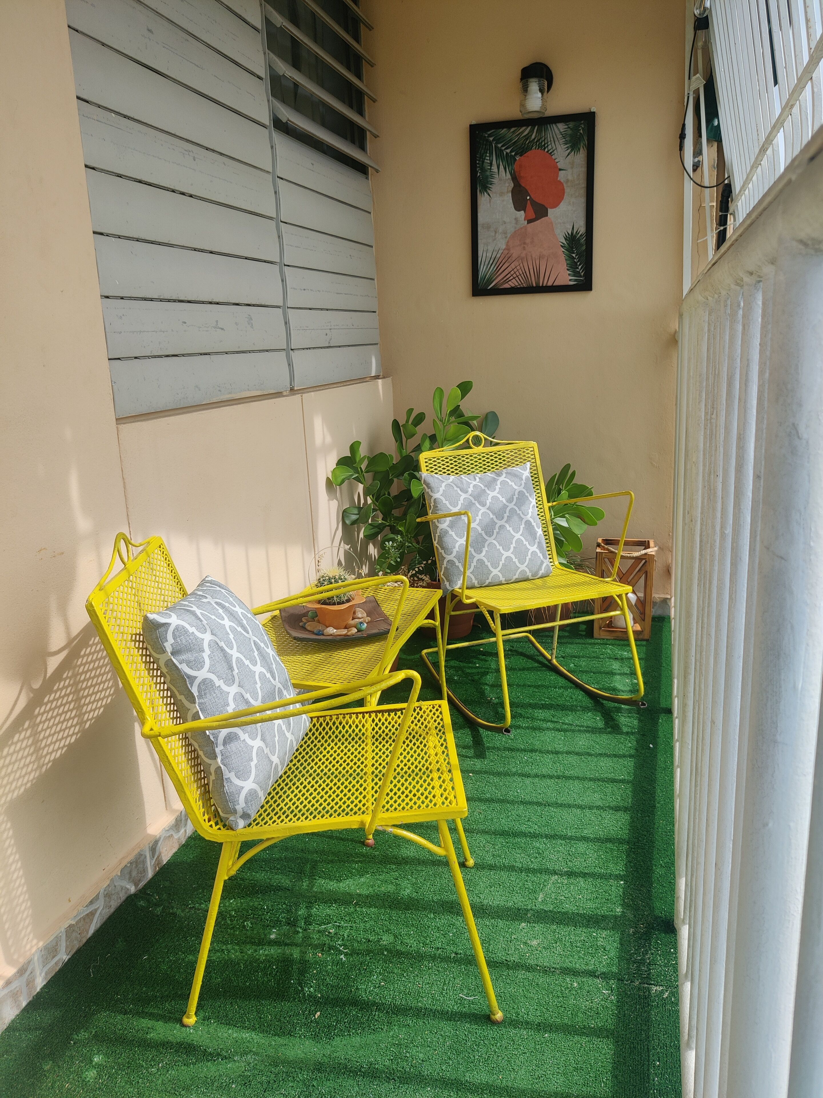 Uma varanda com cadeiras de metal amarelas e uma mesa, grama sintética verde, uma planta em vaso e um quadro na parede.