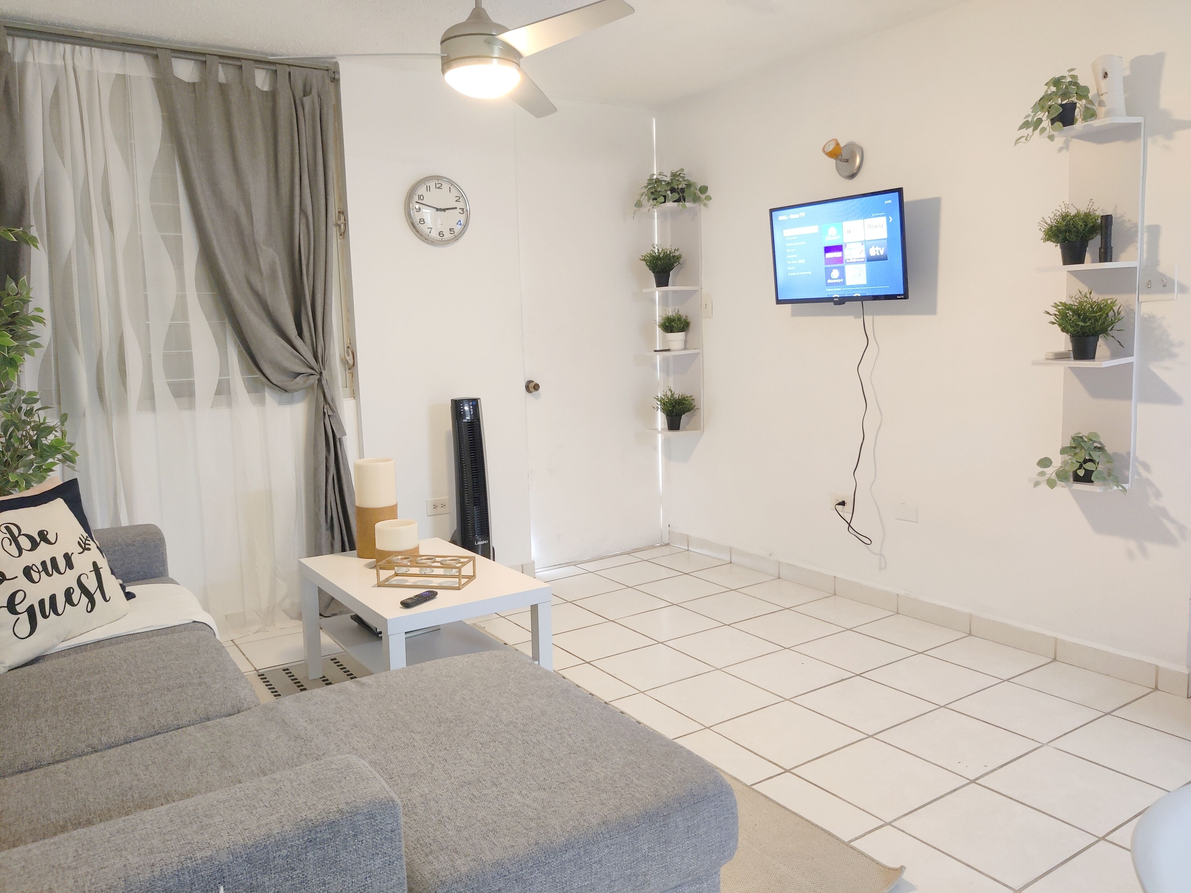 En moderne stue med en grå sofa, et lille bord, et vægmonteret fjernsyn og flere potteplanter.