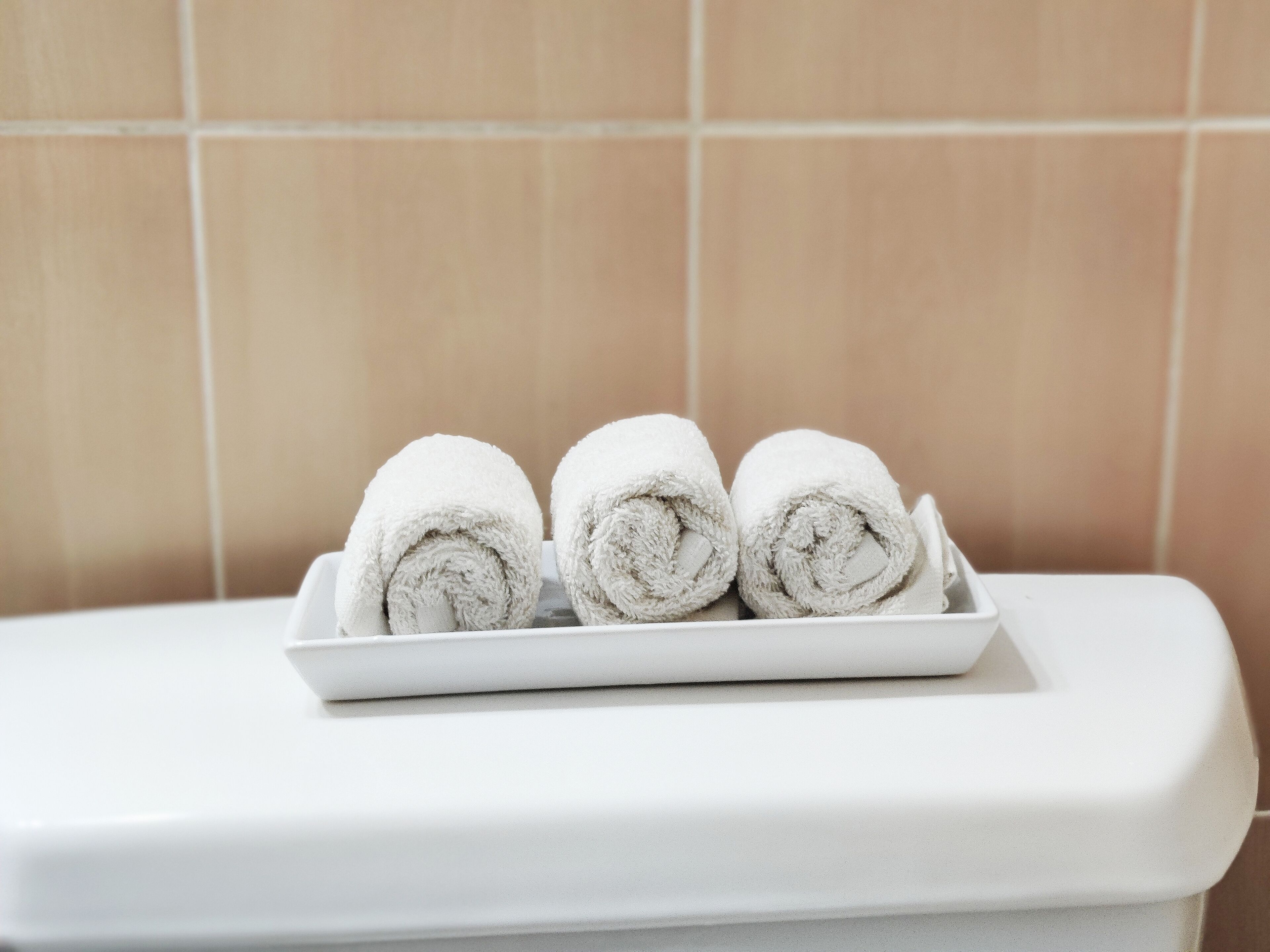 A white tray with three rolled towels on a white surface, likely a bathroom counter, against a tiled wall.