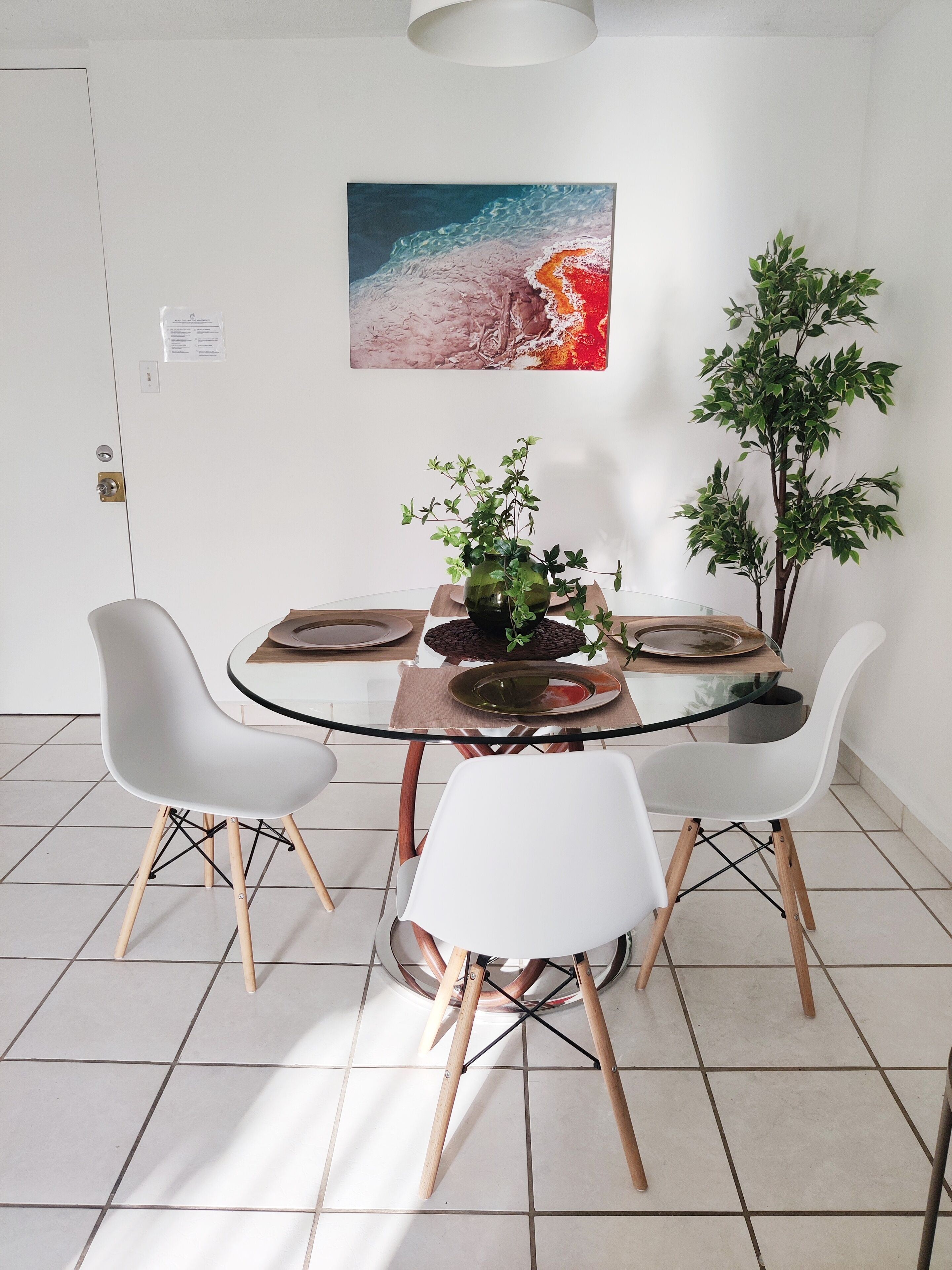 Un coin repas avec une table en verre, des chaises blanches, une plante et un tableau au mur.
