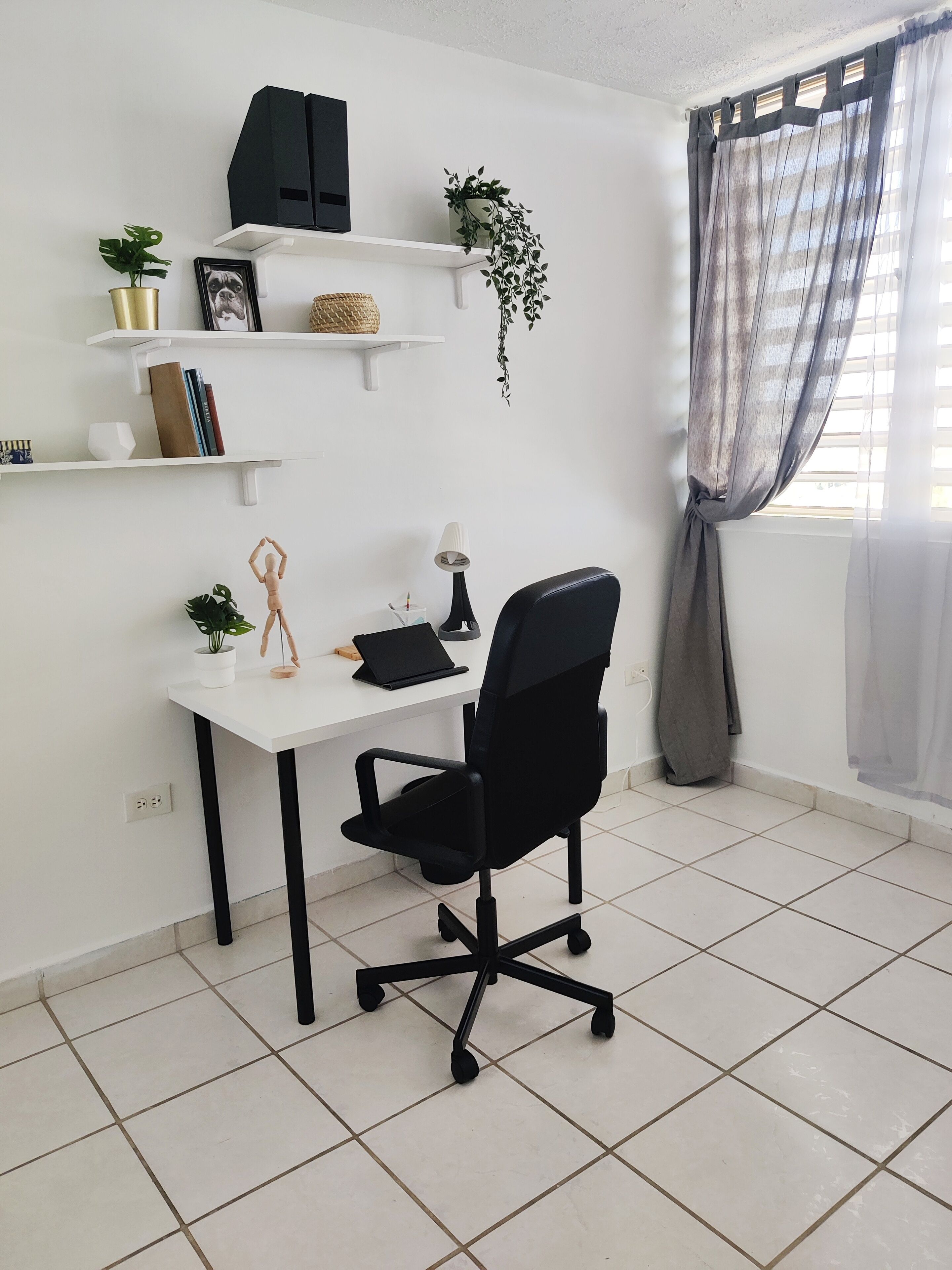 Un espace de bureau propre et moderne, avec un bureau blanc, un fauteuil de bureau noir et des étagères où sont rangés des livres, des plantes et des objets décoratifs.