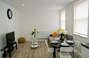 A modern living room with a flat-screen TV, a round glass table with chairs, a sofa with cushions, and a dining table set for two.