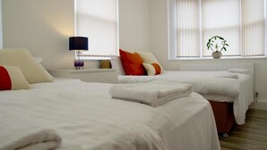 Hotellihuone, jossa on kaksi sänkyä, yöpöytä lampun kanssa, ikkuna, jossa on verhot, ja ruukkukasvi.