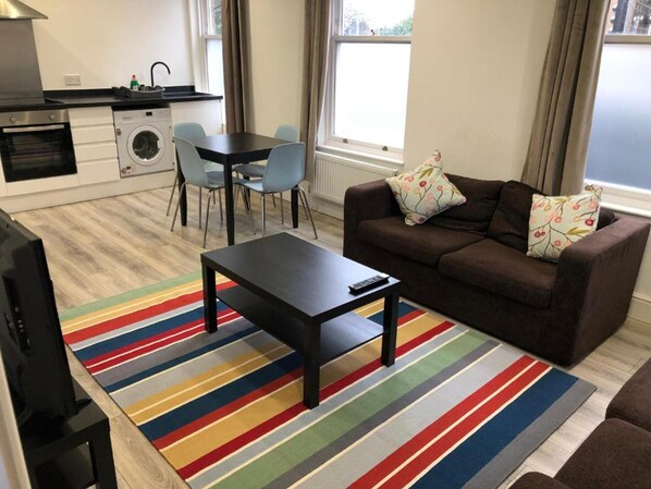 A compact living space with a sofa, a coffee table, a TV, a dining table with chairs, a kitchen area with a washing machine, and a colorful striped rug.