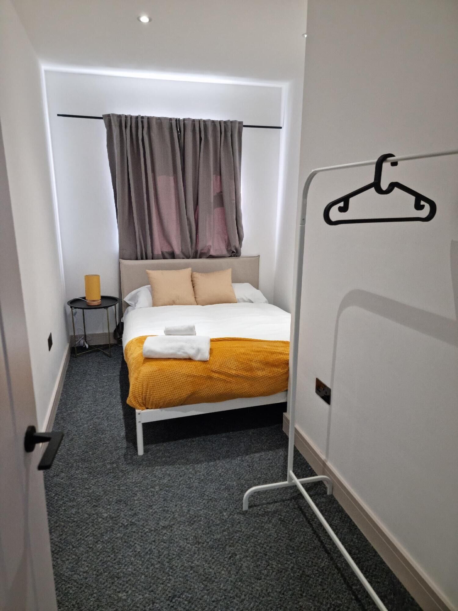 A hotel room with a bed, a bedside table with a lamp, a coat rack, and a door with a hanger symbol.