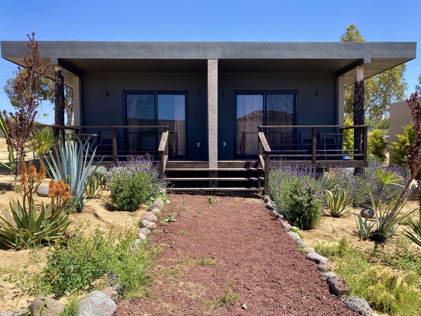 A modern building with a concrete facade, large windows, and a gravel pathway leading to a wooden staircase. - Coquenas del Valle (Valle de Guadalupe)