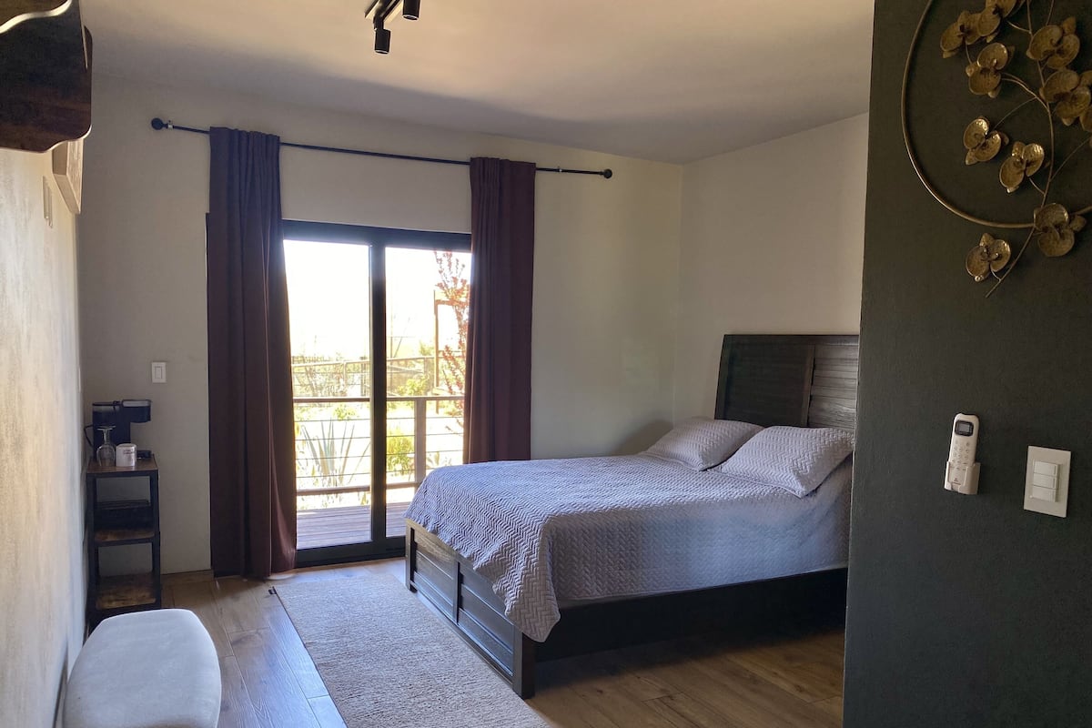 Ein Schlafzimmer mit einem Bett, einer Bank, einer Kaffeemaschine, einem Fenster mit Vorhängen und Blick ins Freie.