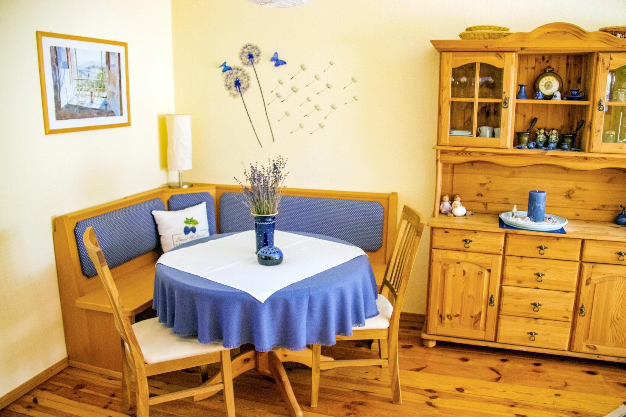 Une pièce comprenant un banc en bois, une table recouverte d’une nappe bleue, un meuble avec des portes en verre, et un mur orné d’un tableau encadré et d’objets décoratifs.