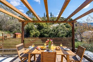 Uma pérgola de madeira com uma mesa posta para refeição, emoldurada por paisagens naturais.