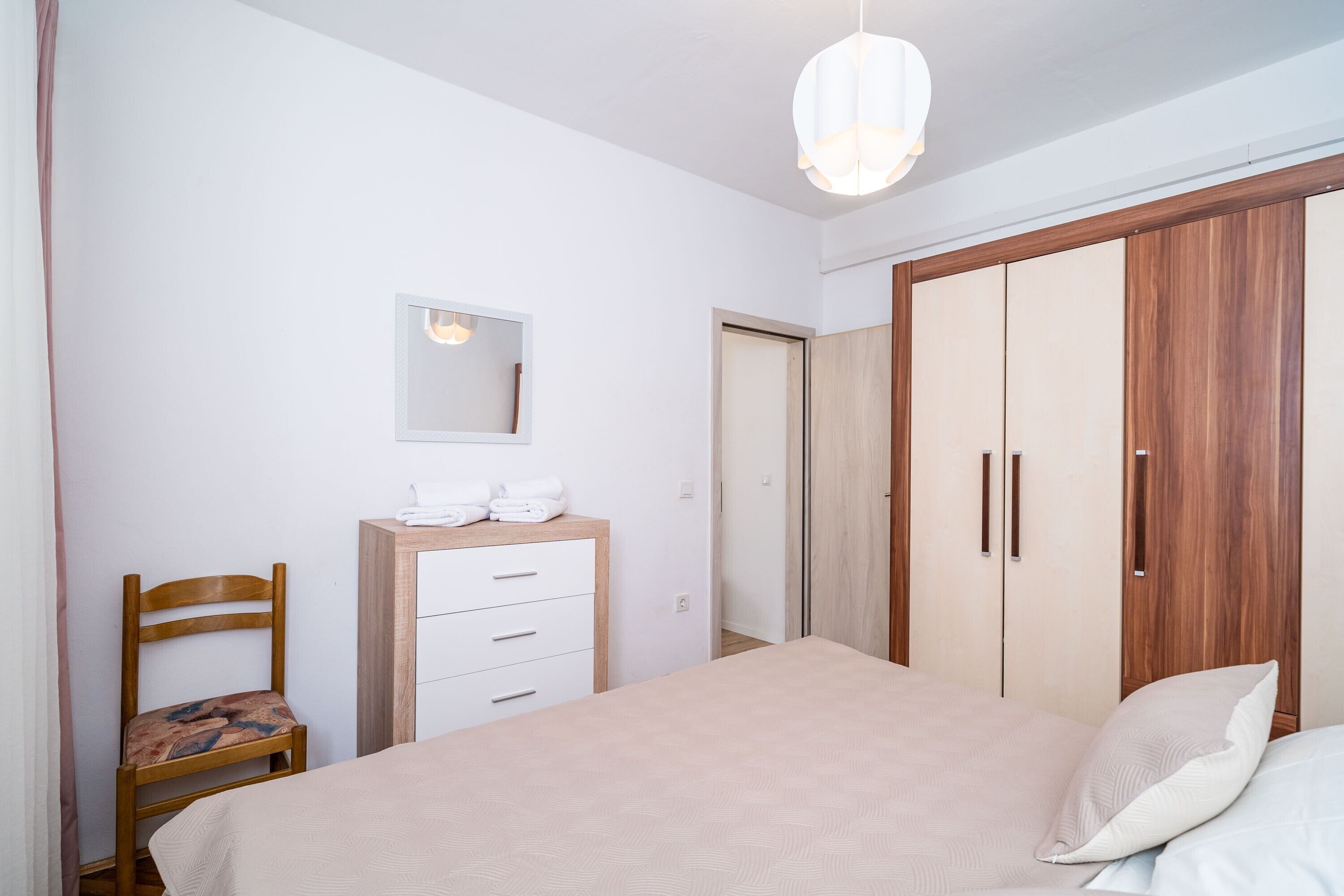 A bedroom with a bed, a wooden wardrobe, a bedside table with towels, a chair, and a hanging light fixture.