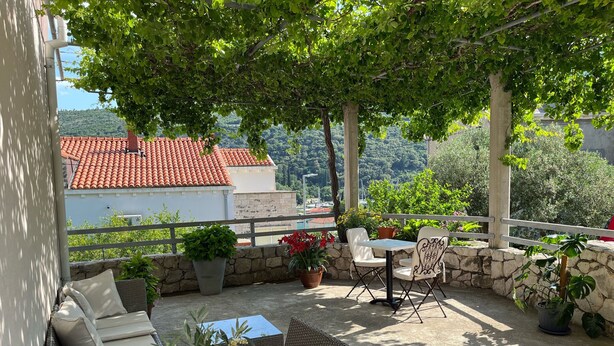A covered outdoor seating area with wicker furniture, potted plants, and a view of a red-tiled building in the background. - Apartment N&I - One Bedroom with Terrace (Dubrovnik)