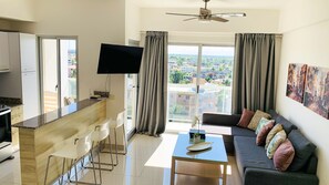A modern apartment with a kitchen, dining area, and living room. It features a flat-screen TV, a sofa with cushions, and a large window with curtains. - Apartamento torre ll (Santo Domingo Este)