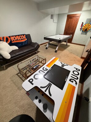 Ein Raum mit Billardtisch, einer Couch mit Broncos Decke, einem Tischtennisplatten und einer Dartscheibe.