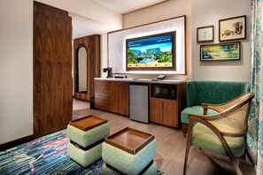 A compact hotel room with a kitchenette, a flat-screen TV displaying a resort, a green armchair, and a colorful rug.