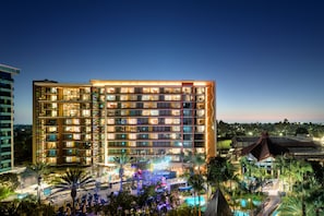 A multi-story hotel with a pool area, palm trees, and a well-lit facade.