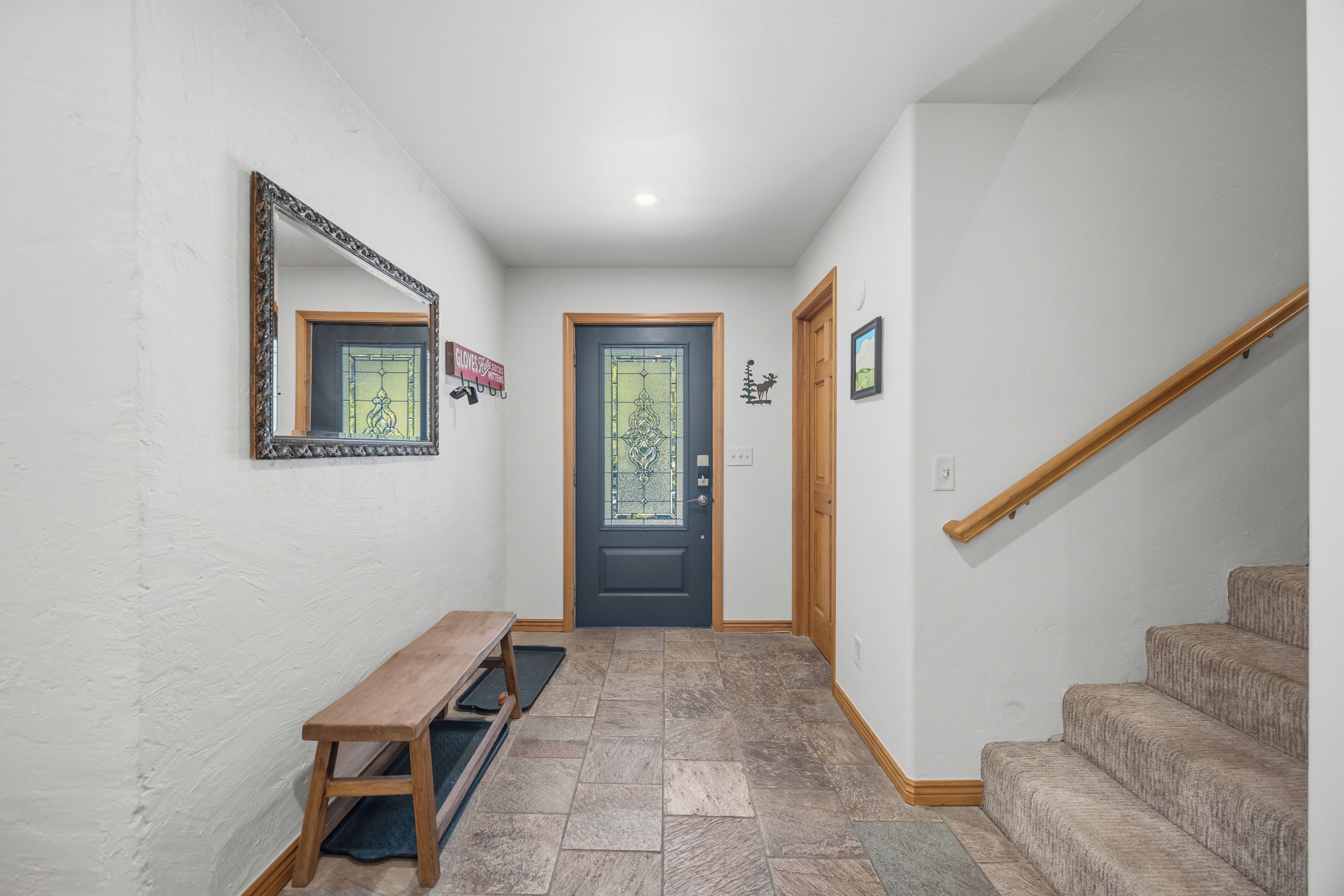 A hallway with a bench, a door with a glass panel, a wooden staircase, and a wall-mounted mirror.